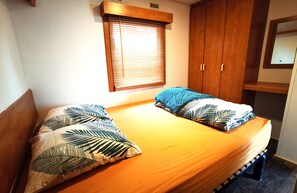 Un lit double avec des oreillers à motifs de feuilles, une tête de lit en bois, une fenêtre avec des stores et une armoire en bois dotée d’un miroir.