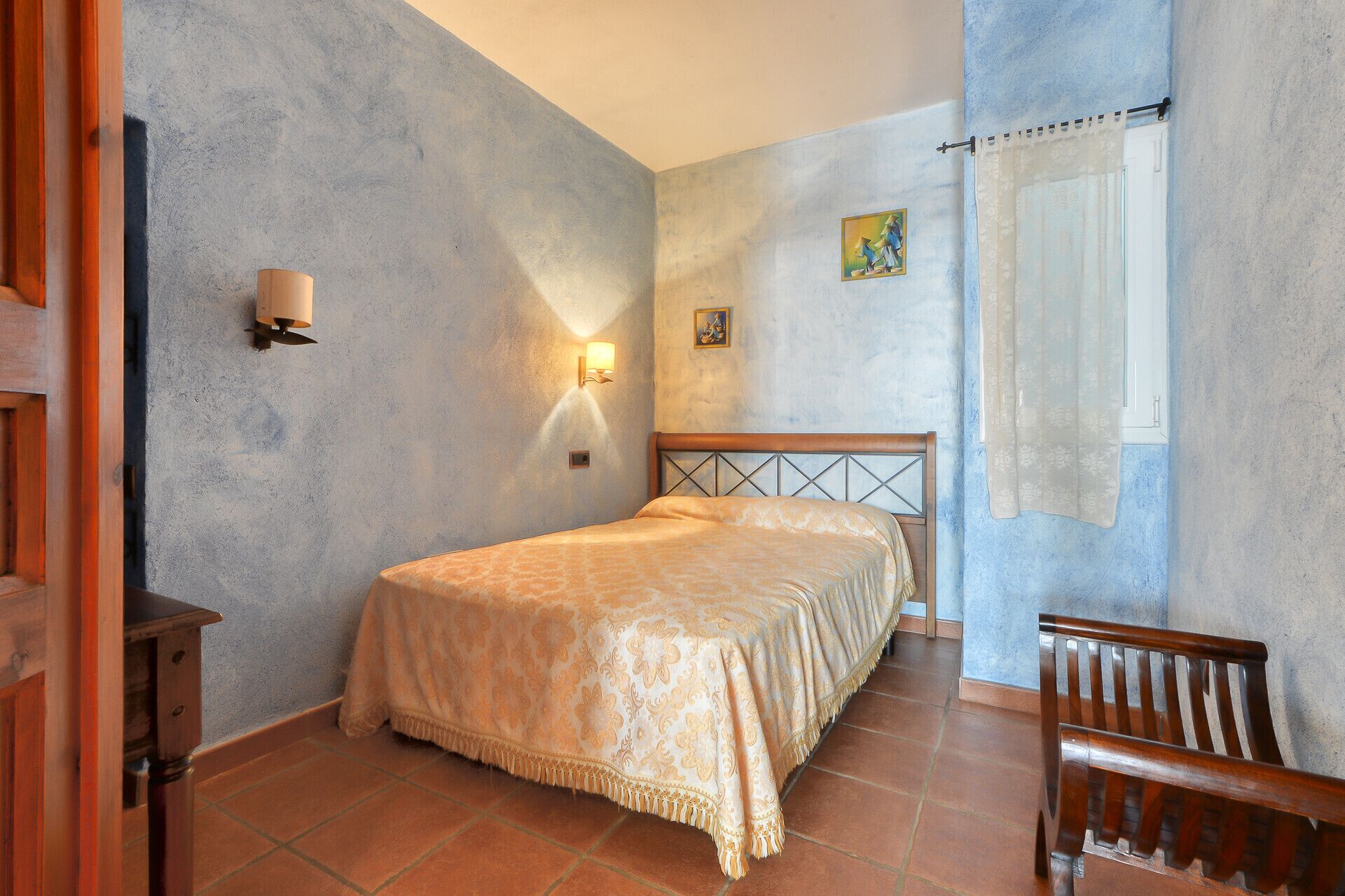 A bedroom with a bed, a nightstand, a wall-mounted lamp, a window with a sheer curtain, and a small painting on the wall.