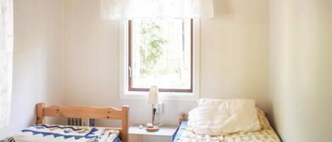 A small, well-lit bedroom with two single beds, a window with a view of greenery, and a bedside table with a lamp.