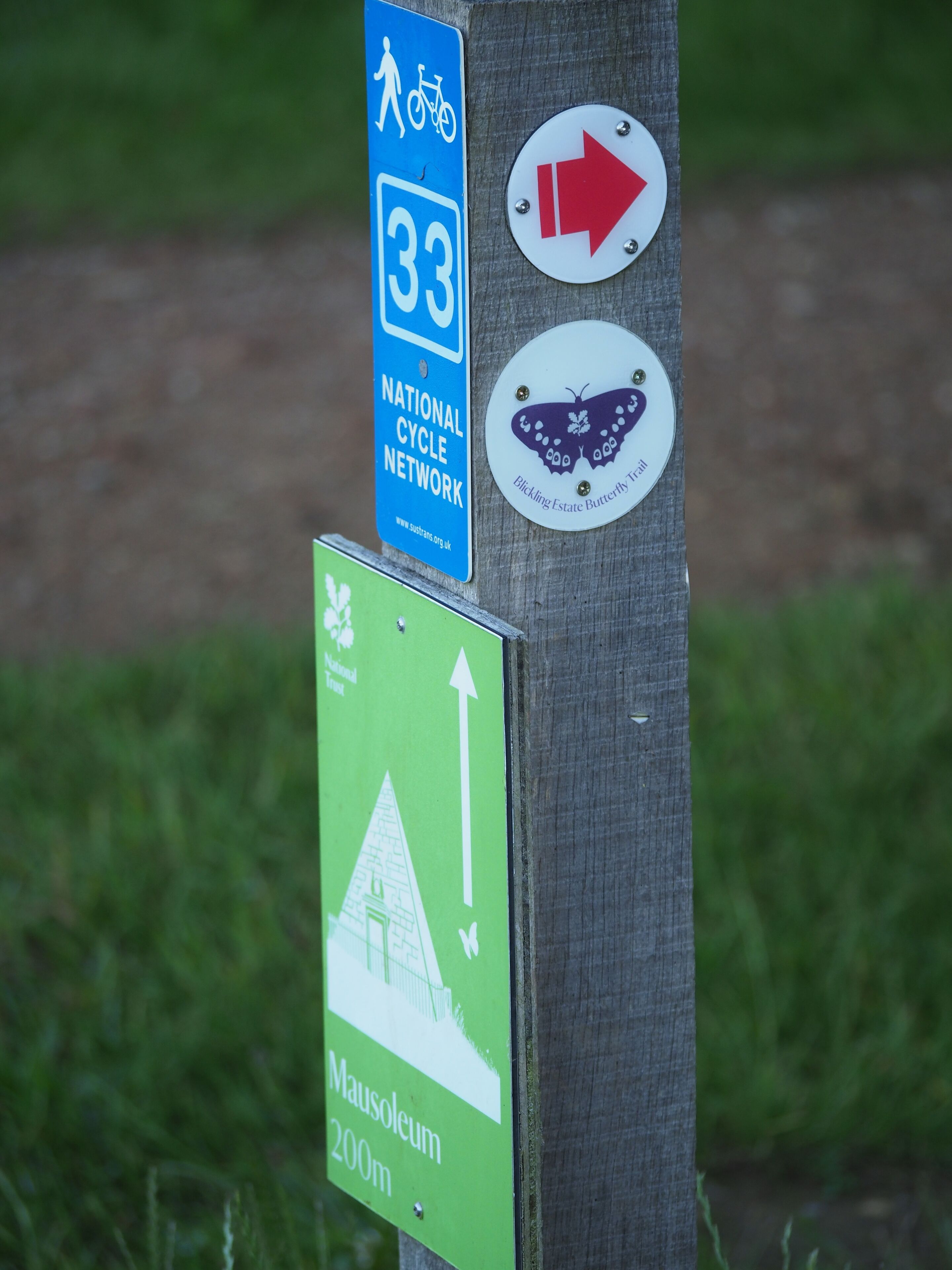 A wooden post with multiple signs: a blue sign for the National Cycle Network route 33, a red arrow sign, a purple butterfly sign for Bickling Estate Butterfly Trail, and a green sign for Mausoleum 200m.
