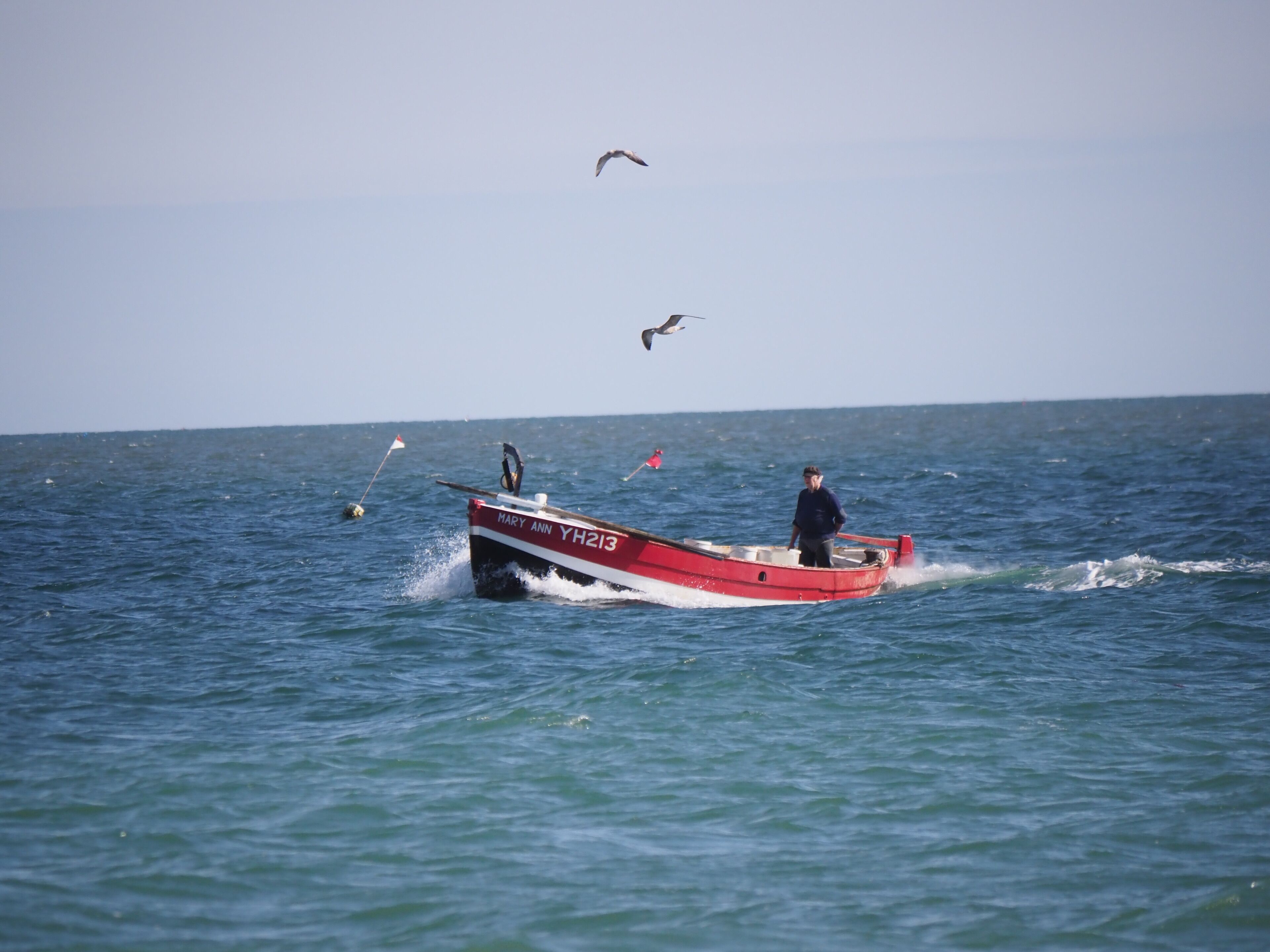 Un petit bateau rouge et blanc, baptisé « MARY ANN », flotte sur l’eau, et une personne est à la barre.