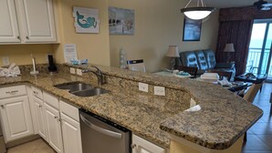 A kitchen with granite countertops, white cabinets, a dishwasher, and a sink. There are decorative wall art pieces and a view of the ocean through a window. - 25 ft. Balcony Oceanfront ! Incredible views !MONTHLY WINTER RENTAL AVAILABLE ! (Myrtle Beach)