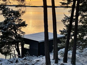A cabin with a flat roof, surrounded by snow-covered trees and a body of water in the background. - Lakeside chalet with spectacular views and exclusive private location  (Alingsås NV)