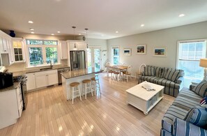 A modern kitchen with white cabinets, a central island with stools, a dining area with a striped sofa, and a large window providing natural light.