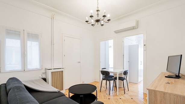 A modern living room with a sofa, a dining table with chairs, a TV, and a chandelier.