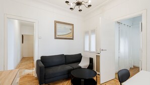 A modern living room with a dark sofa, a round coffee table, and a white table. There is a painting on the wall and a door leading to another room.