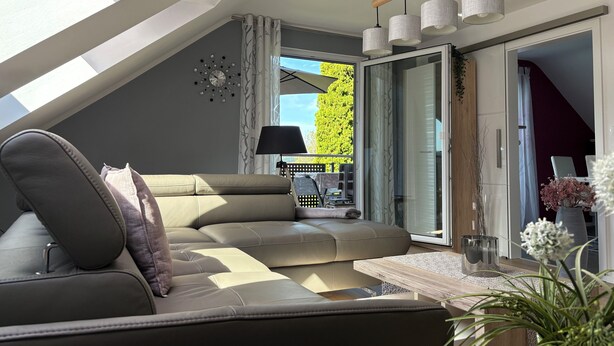 A modern living room with a sectional sofa, a side table, and a lamp. There is a sliding glass door leading to an outdoor area with a patio set. - Modern apartment in Schauenstein with loggia and balcony **** (Schauenstein)