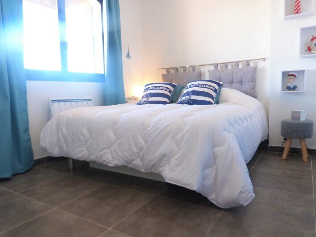 Ein ordentlich bezogenes Bett mit weißer Daunendecke, zwei Kissen mit Meeresmotiven, ein kleiner Nachttisch und ein an der Wand befestigtes Regal mit einer Figur.