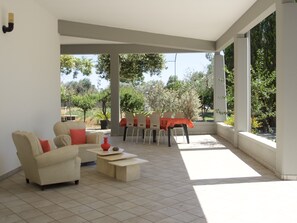 Un espace extérieur aménagé, abrité et doté d’un canapé, de chaises et d’une table, entouré de grandes fenêtres et de verdure.