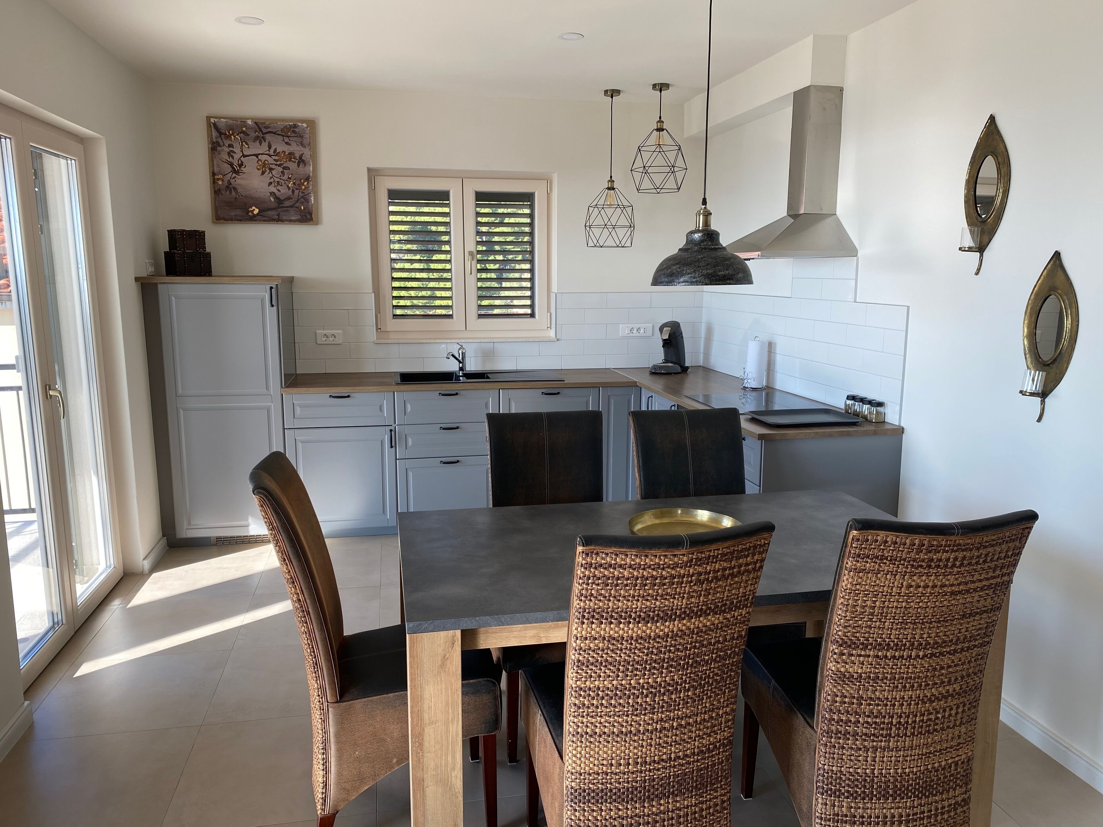 Una cucina moderna con zona pranzo, caratterizzata da un'ampia finestra, lampade a sospensione e uno specchio decorativo.