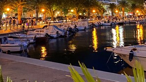 Ein Yachthafen mit vertäuten Booten, eine Uferpromenade und eine gebirgige Kulisse.