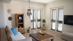 Ein Wohnzimmer mit einer Couch, Couchtisch und Fernseher. Zwei Fenster mit Jalousien, ein Bücherregal und eine Pendelleuchte sind ebenfalls vorhanden.