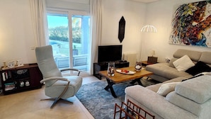 Un salon moderne avec un canapé gris, un fauteuil en cuir, une table basse en bois et un téléviseur. On y trouve une bibliothèque remplie de livres et d’objets décoratifs, un tableau accroché au mur et une vue à travers la porte vitrée.