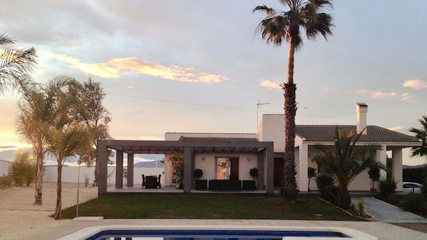 Une maison moderne avec une piscine, entourée de palmiers et sous un ciel dégagé.