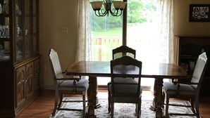 Une salle à manger avec une table en bois, quatre chaises, un lustre et une vue sur une terrasse, visible à travers une grande fenêtre.