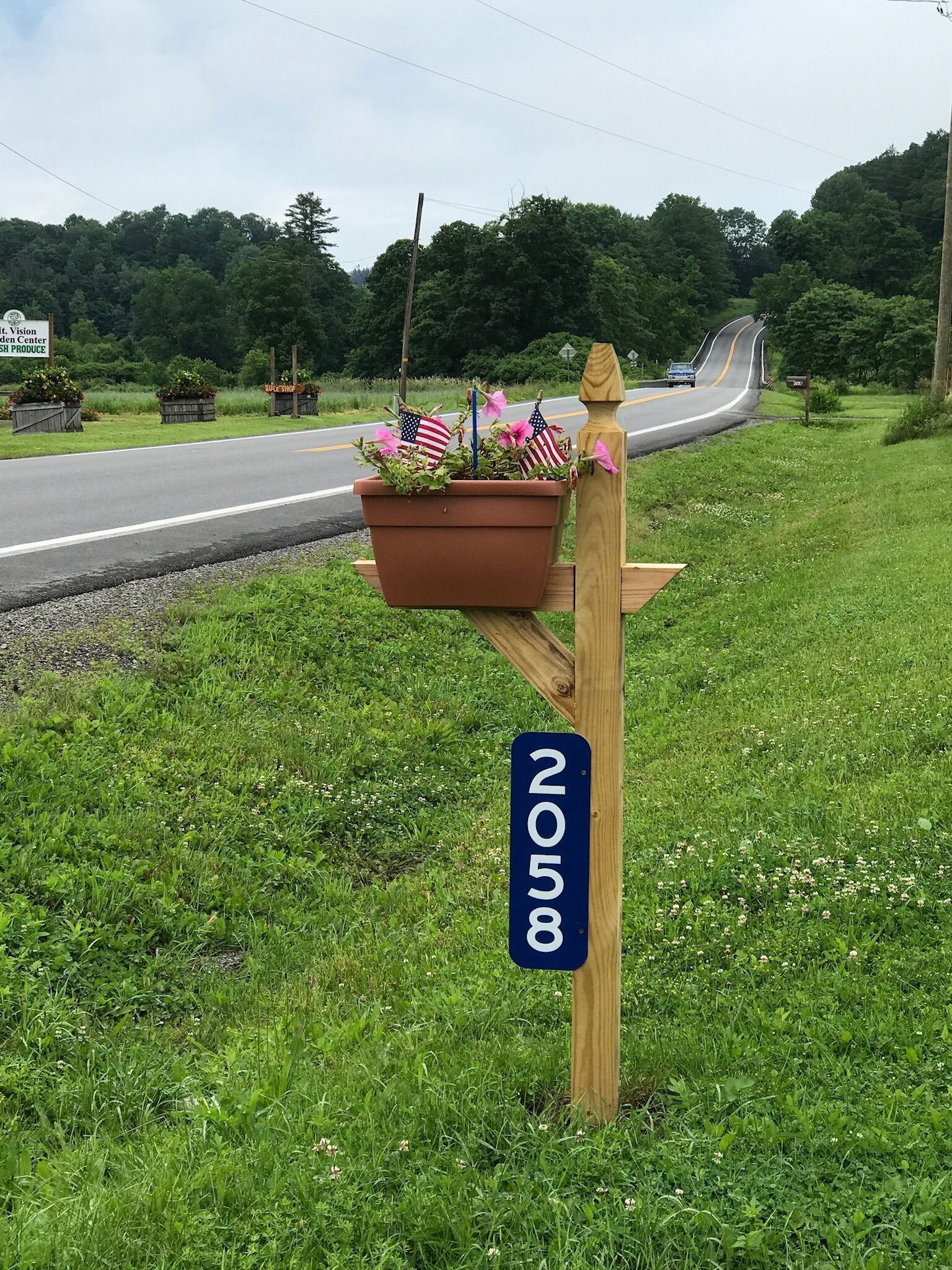 A rural roadside mailbox with a wooden post, a blue sign with the number '8518', and a planter with flowers and an American flag.