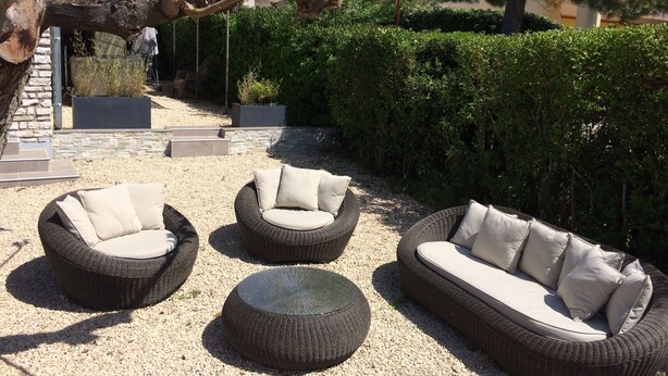 Un espace patio aménagé avec des meubles en osier, notamment un canapé et deux fauteuils, entouré de gravier et de verdure.