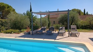 Un espace aménagé au bord de la piscine, avec des chaises longues et une pergola, entouré de verdure et bordé d’un bâtiment dont le toit est recouvert de tuiles.