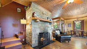 A cozy living room with a stone fireplace, wooden flooring, a leather sofa, and a dining area with a chandelier. - Cabin near BR/Boone, fire pit, WIFI, secluded, nearby hikes, and VIEWS!! (Blowing Rock)