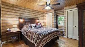 A log cabin bedroom with a bed, a nightstand, a ceiling fan, and a window with shutters. - Cabin near BR/Boone, fire pit, WIFI, secluded, nearby hikes, and VIEWS!! (Blowing Rock)