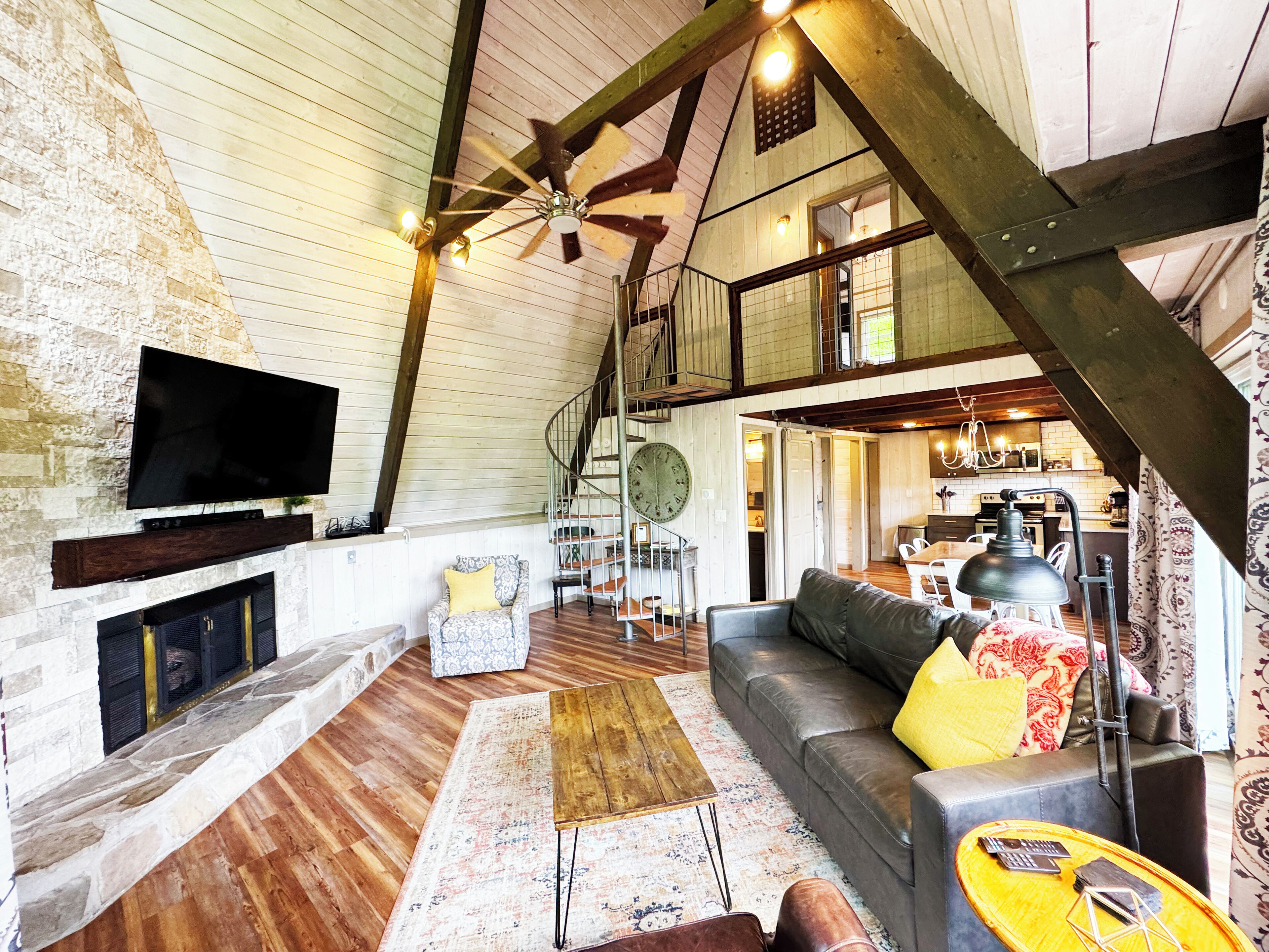 A spacious living area with a wooden ceiling, a large TV mounted on the wall, a staircase leading to an upper level, and a cozy seating arrangement with a sofa and a coffee table.