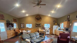 A spacious living room with a gray sofa, a wooden coffee table, and a red armchair. There is a kitchen area with wooden cabinets and a stove. - Chippewa View Heights ~ breathtaking views and a peaceful getaway! (Durand)