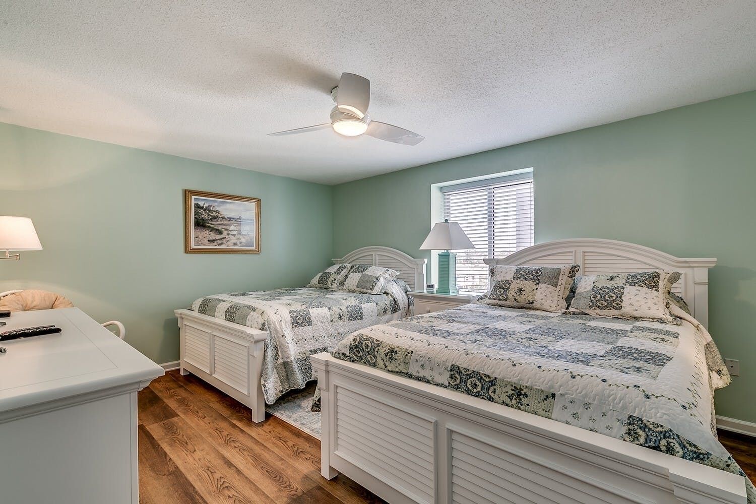A bedroom with two beds, a ceiling fan, a window with blinds, and a painting on the wall.