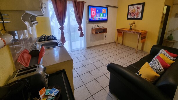 Un espacio habitable compacto con kitchenette, un televisor de pantalla plana montado en la pared, un sofá con cojines y una pequeña mesa con un florero.