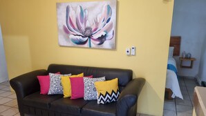 Una sala de estar con un sofá de cuero negro, cojines coloridos, un cuadro grande con motivos florales en la pared y una mesita de noche con un jarrón.