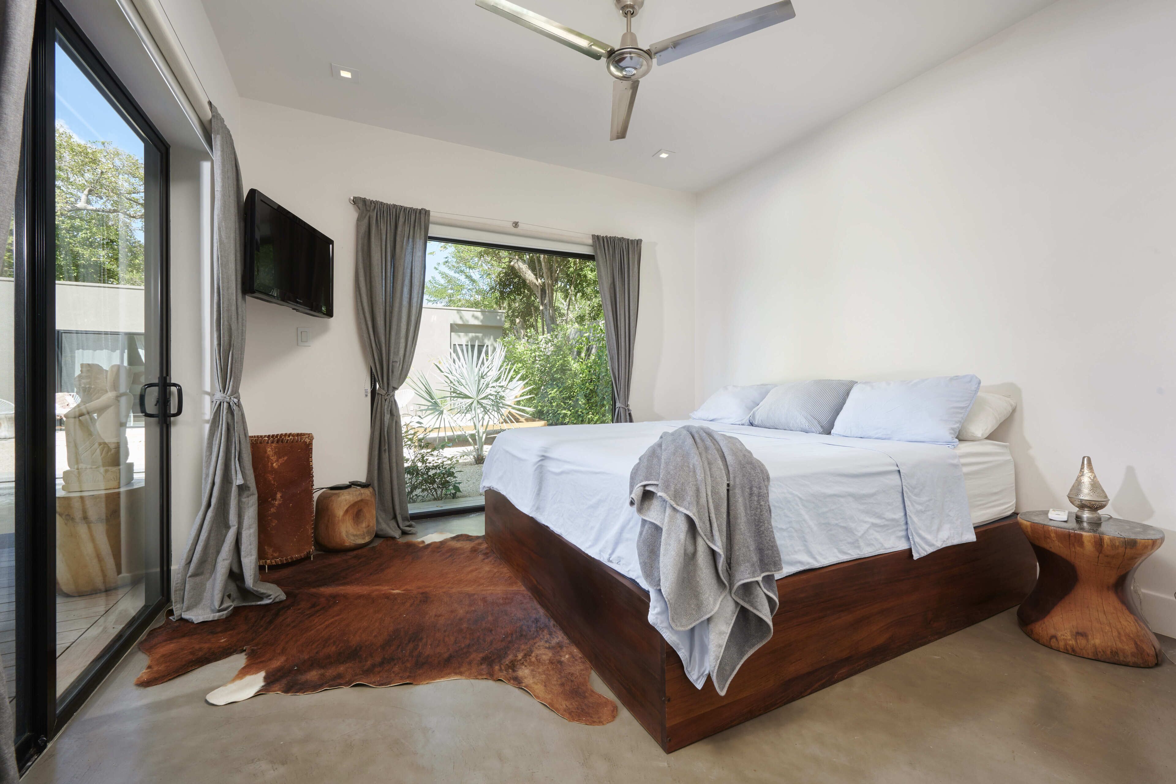 Un dormitorio con una cama grande, ventilador de techo, televisor y vistas a zonas verdes.
