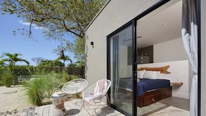 Ein modernes Schlafzimmer mit einer großen Glasschiebetür, einem hölzernen Bett und zwei weißen Stühlen auf einer Holzdielen-Terrasse.