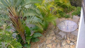 Un patio con suelo de piedra, una mesa redonda con sillas y diversas plantas tropicales.