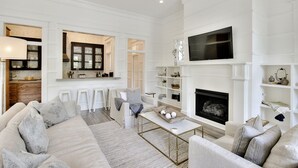 A modern living room with a sofa, coffee table, and fireplace. There is a kitchen area with bar stools and a dining table. - Luxury Living Awaits: 5-Bed Retreat with Pool, Bikes, and Golf Cart in WaterColor! (Santa Rosa Beach)