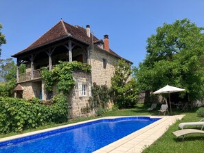Et stenhus med swimmingpool, omgivet af grøn vegetation og en klar himmel.