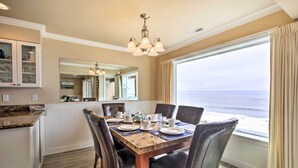 Un coin repas avec une table en bois dressée pour quatre personnes, entourée de chaises, d’un lustre et d’une grande fenêtre donnant sur l’océan.
