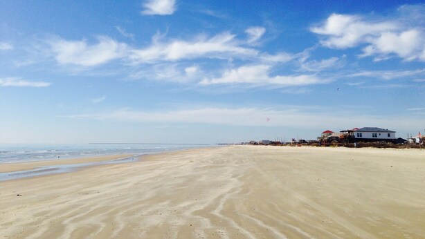 Une vaste plage de sable fin, avec un ciel dégagé et quelques nuages. On aperçoit des bâtiments à l’arrière-plan, près du bord de l’eau.