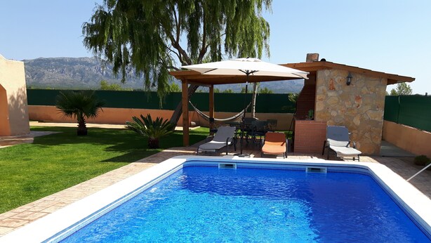 Una piscina con una pérgola y sillas de descanso, rodeada por un césped cuidado y un muro de piedra.