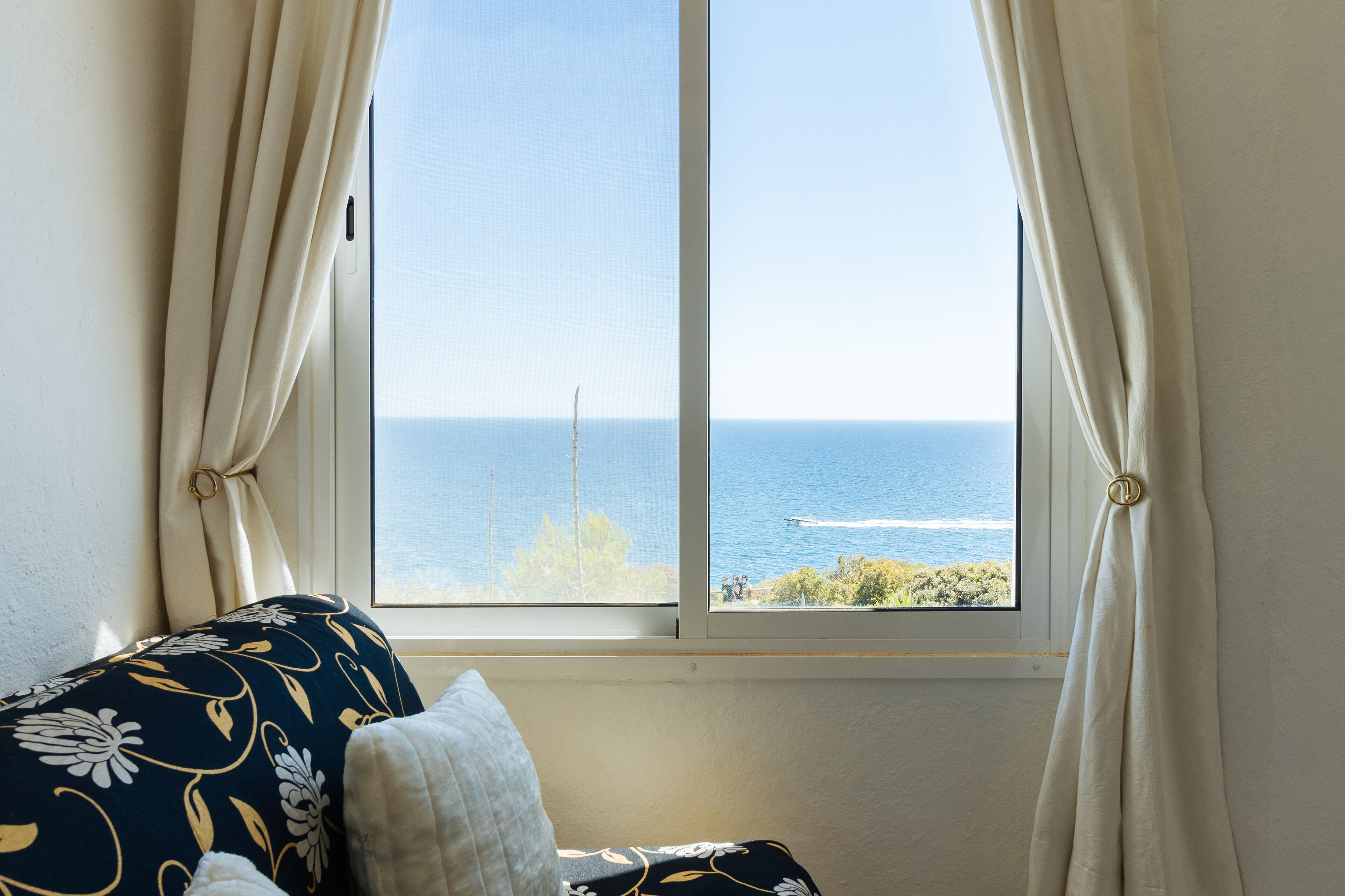 Une chambre avec vue sur la mer, visible par une fenêtre, un lit agrémenté d’un coussin à motifs et un rideau retenu.