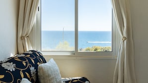 Een kamer met uitzicht op de zee door een raam, een bed met een kussen met een patroon, en een gordijn dat is opgebonden.
