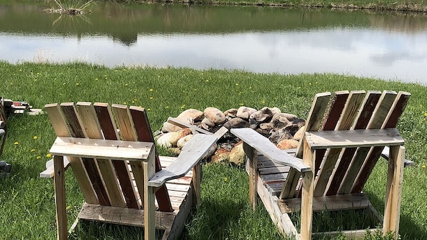 Dos sillas Adirondack mirando a un brasero, con un lago y una colina cubierta de pasto al fondo.
