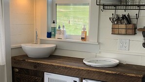 Una cocina compacta con un lavamanos blanco, un microondas y un refrigerador. Hay una ventana sobre el lavamanos, y una repisa con utensilios de cocina.