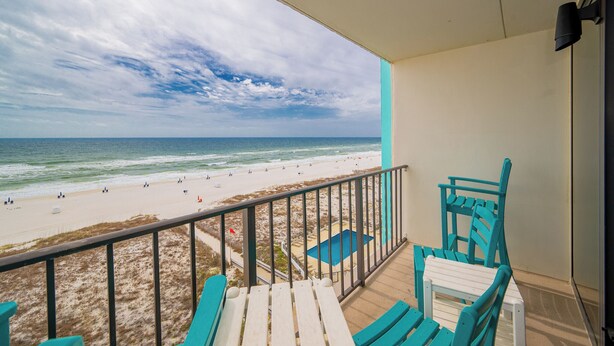 A balcony with a view of a beach and ocean. There are turquoise chairs and a small pool.
