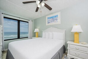 Une chambre avec un grand lit, un ventilateur de plafond, une fenêtre donnant sur l’océan et un tableau accroché au mur.