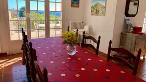 A dining area with a red tablecloth, wooden chairs, a vase with yellow flowers, and a view of the outdoors through large windows. - Villa 12 persons with heated swimming pool, sea view  (Saint-Raphaël)