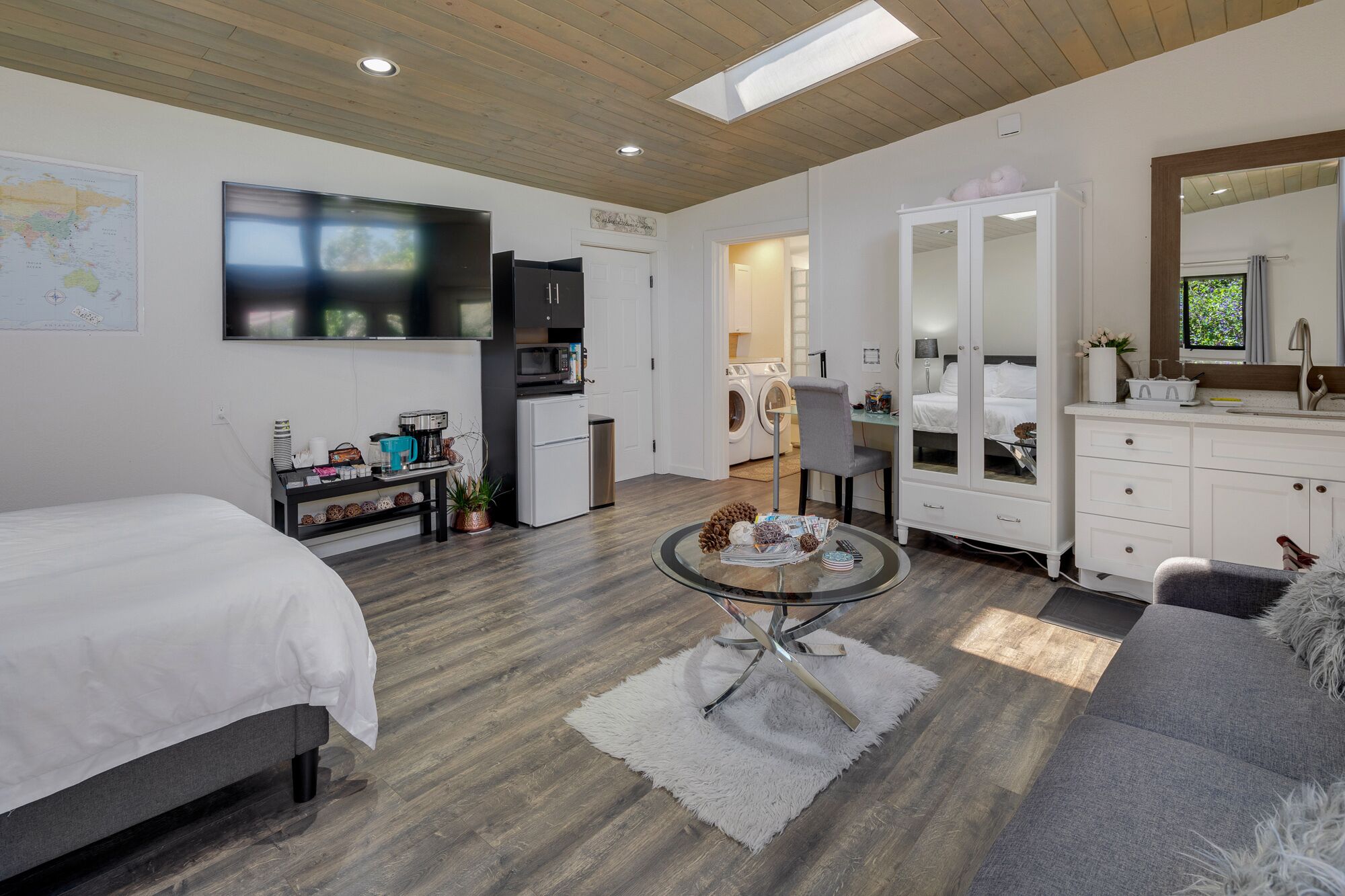 A modern hotel room with a bed, a sofa, a dining table, a kitchen area, and a bathroom with a large mirror.