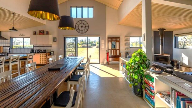 A spacious kitchen with a large wooden table and chairs, a clock on the wall, and a view of the outdoors through large windows. - Abercrombie Ridge - Lux BARN and Spa Retreat - Pet-friendly (Curraweela)