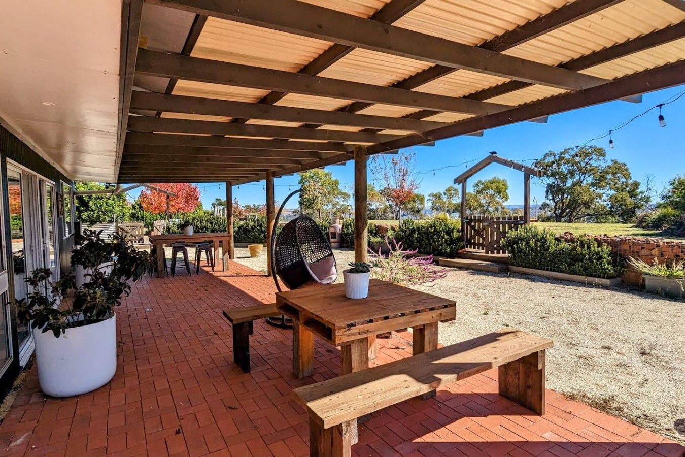 An outdoor patio area with a wooden table and benches, surrounded by potted plants and a gravel path.