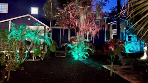 Une maison à l’extérieur d’un bleu foncé, avec une fenêtre visible et une porte. Des plantes et des arbres entourent la maison, et certains sont illuminés de lumière verte.