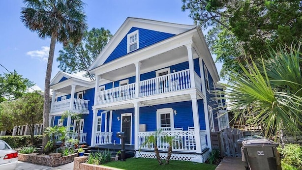 Une maison à deux étages, à l’extérieur bleu et aux ornements blancs, dotée d’un porche avec des balustrades blanches et d’un petit jardin devant, agrémenté de plantes et d’une terrasse en bois.