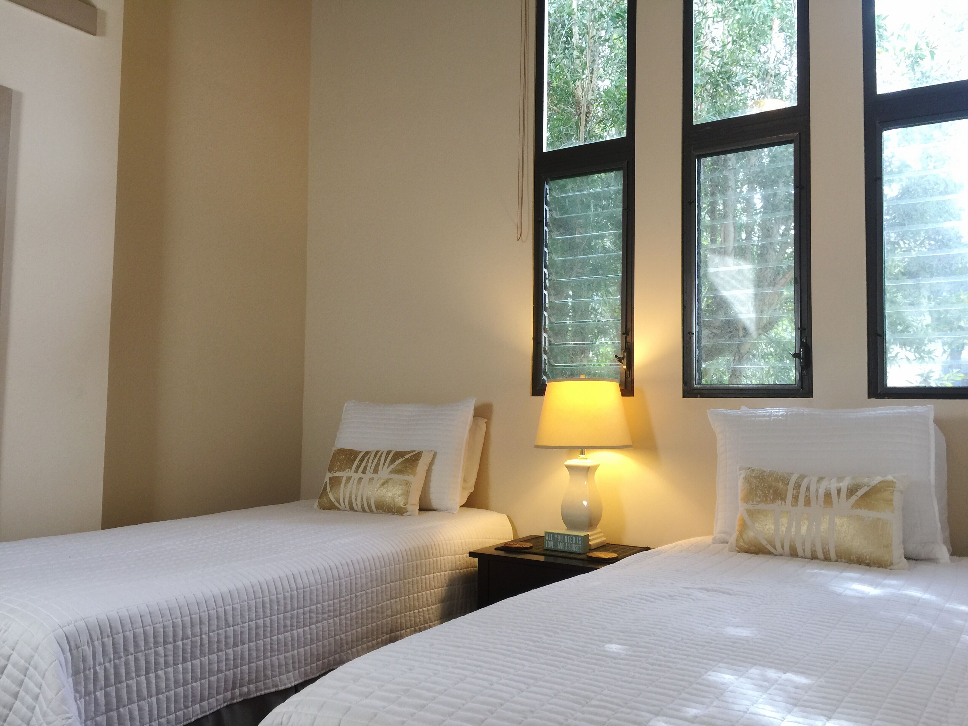 A hotel room with two beds, a nightstand with a lamp, and large windows with a view of trees.